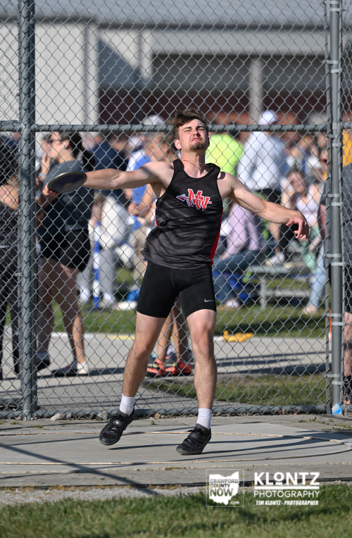 Male discus thrower in a black singlet releases a discus during a track and field event, behind a chain-link fence with spectators.