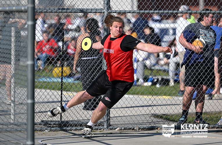 Male player in a red pinnie throws a yellow frisbee during a game behind a chain-link fence with spectators in the background.