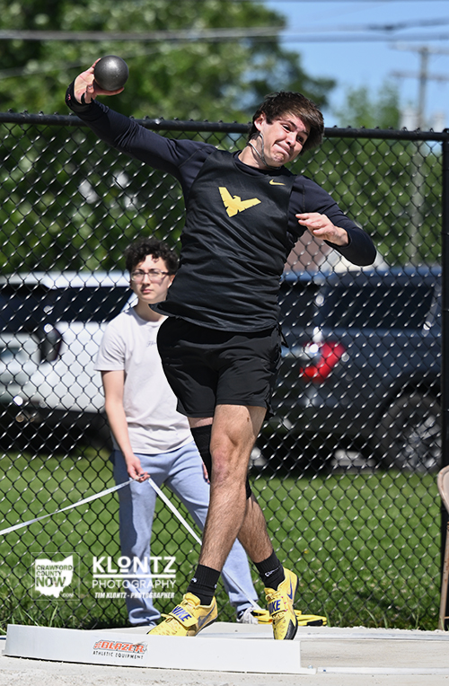 Man in black athletic gear mid-throw during a shot put on an outdoor competition platform, with a spectator watching behind a chain-link fence.