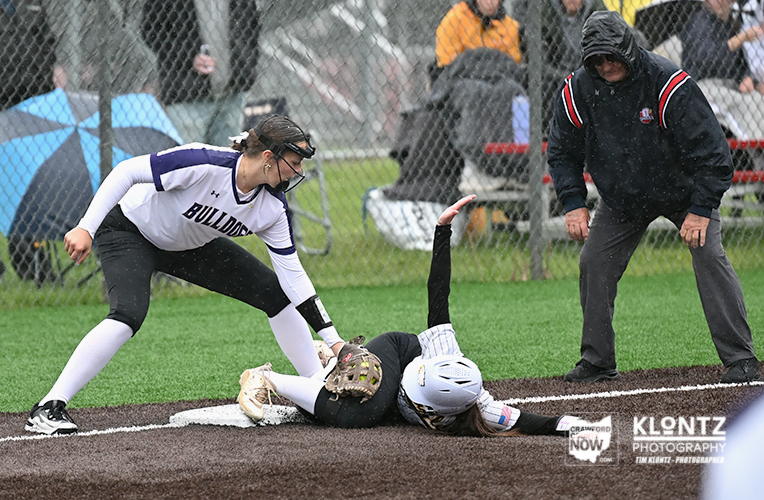 Softball: fielder in purple and white tags a sliding baserunner at a clover-shaped infield base while a coach watches nearby on a rain-slick field behind a chain-link fence.