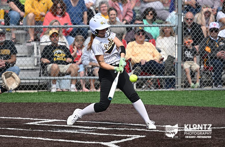 Female softball player in white and black uniform swinging at a pitched ball with spectators behind a chain-link fence.