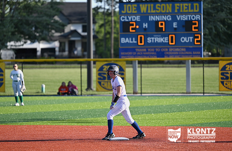 Baseball player in a white uniform runs the bases on a sunlit field with a blue scoreboard behind him at Joe Wilson Field.
