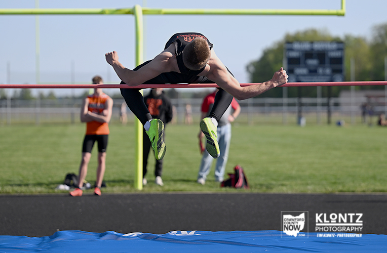 Male high jumper clears the bar mid-air during a track and field event, green shoes visible above the blue pit.
