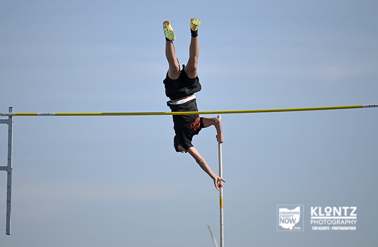Pole vaulter inverted mid‑flight, clearing the bar against a bright blue sky.