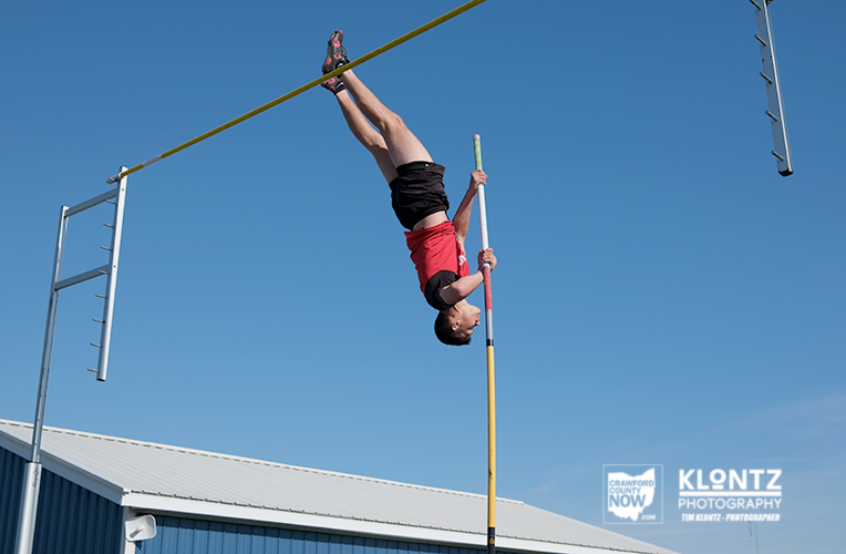 Pole vaulter in a red shirt and black shorts upside down, clearing a yellow crossbar outdoors against a clear blue sky.