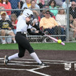 Female softball player in a white striped jersey swings pink bat at a pitched ball near home plate, with spectators behind a chain-link fence.