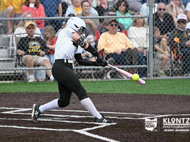 Female softball player in a white striped jersey swings pink bat at a pitched ball near home plate, with spectators behind a chain-link fence.