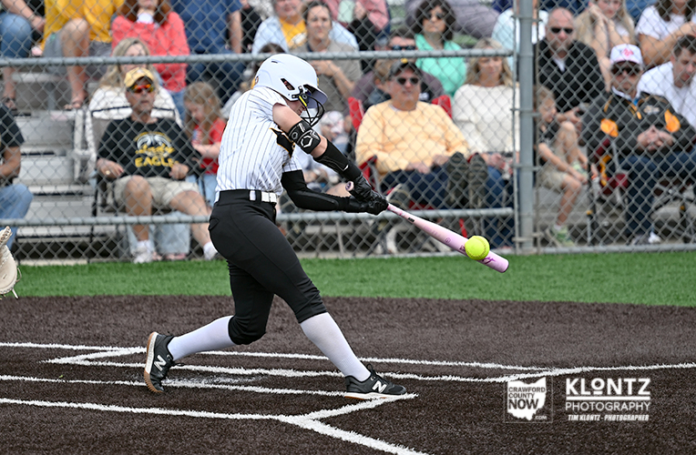 Female softball player in a white striped jersey swings pink bat at a pitched ball near home plate, with spectators behind a chain-link fence.