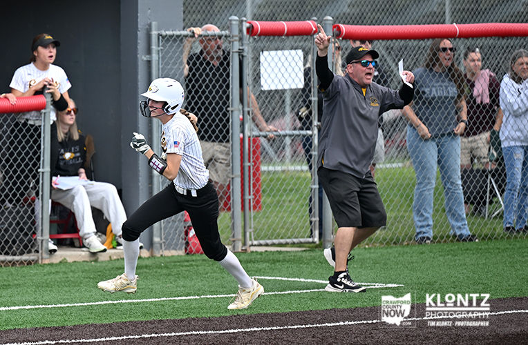 Female softball runner in a white uniform and helmet sprints toward first base as an official in a gray shirt raises arms on the sideline; spectators watch behind a chain-link fence.