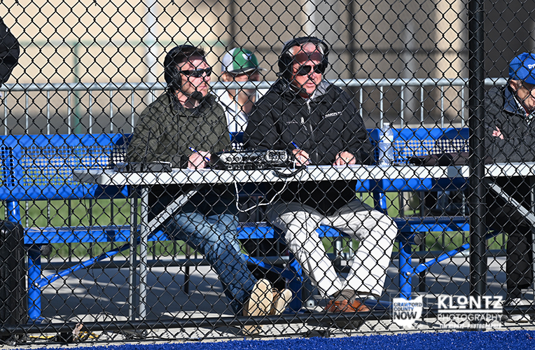 Two men wearing sunglasses sit at a table with radio equipment behind a chain‑link fence at a baseball field.