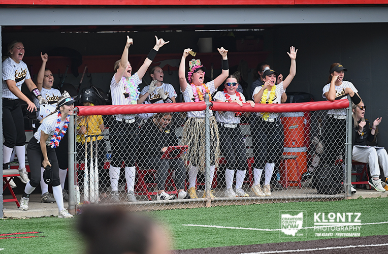 Softball team in a dugout cheering with arms raised, wearing white jerseys and black pants, decorated with leis, behind a chain-link fence on a grassy field.