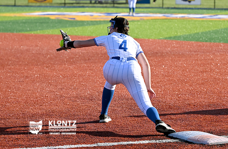 Female softball player in a white pinstripe uniform lunges to catch the ball at a base on an orange infield.