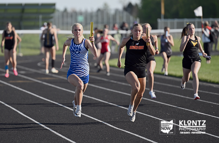 Women’s relay runners sprinting on a track, baton handoffs under a sunny day, with teammates in blue, black, and red uniforms ahead of the pack.