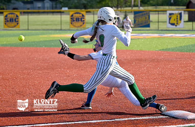 Female softball player in white with green accents runs toward first base as a defender in blue reaches for the ball on the red dirt infield.