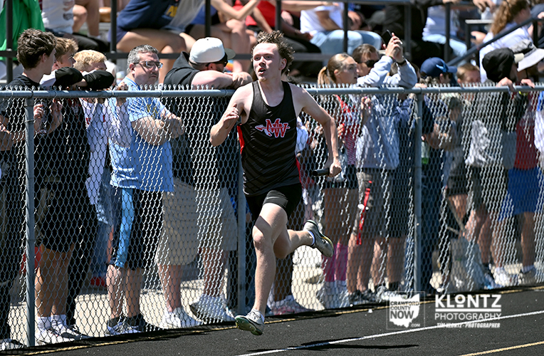 Male sprinter in a black singlet crosses the finish line as cheering spectators stand behind a chain-link fence.