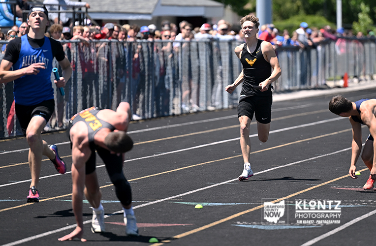 Male sprinters race on an outdoor track near the finish line, with a cheering crowd behind a chain-link fence.