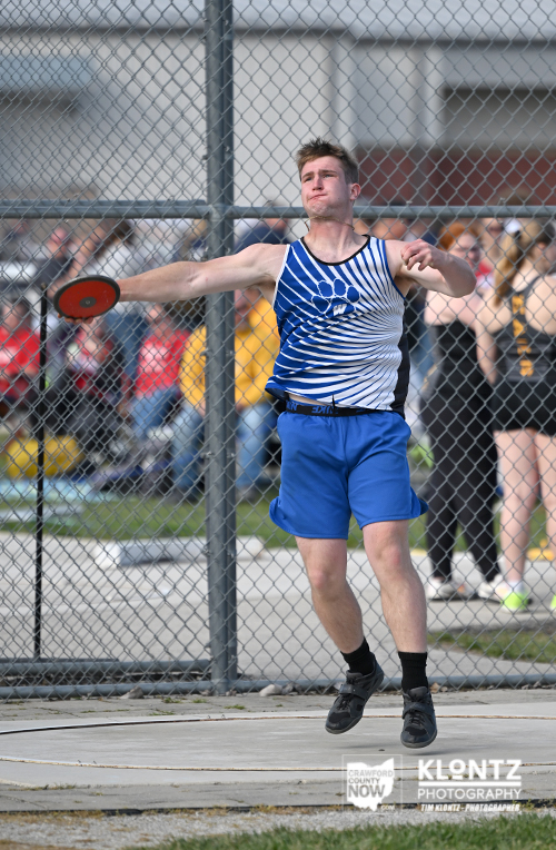 Male athlete in blue shorts and a blue-and-white striped tank top throws a discus during a track and field event behind a chain-link fence.