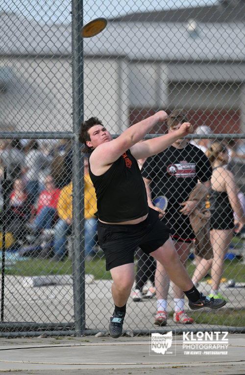 Athlete in a black tank top and shorts throws a discus at an outdoor track and field event behind a chain-link fence.