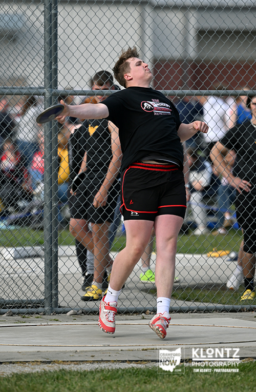 Athlete in a black shirt and red shorts mid-throw of a discus at a track and field meet, with a chain-link fence and spectators in the background.