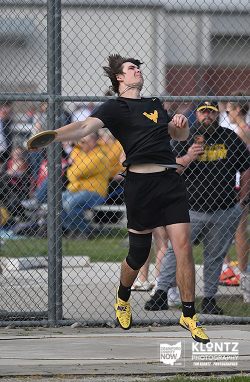 Athlete in black uniform mid-throw of a discus, behind a chain-link fence at a track meet, wearing bright yellow shoes.