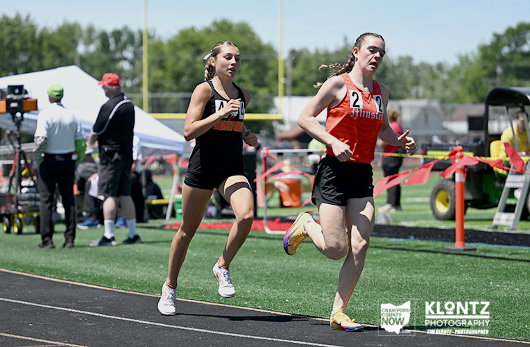 Two young female sprinters race on a track, numbers 4 and 2 visible, with officials and equipment in the background on a sunny day.