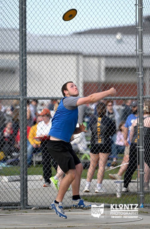 Athlete in a blue shirt throws a discus inside a chain-link fence at an outdoor track meet.