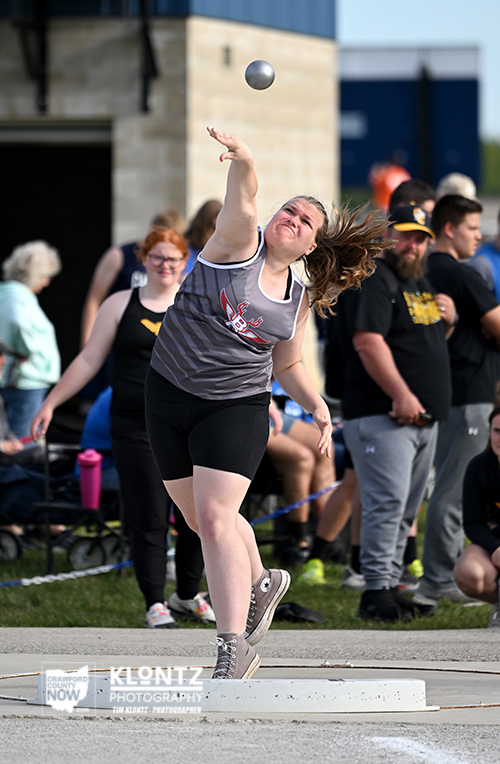 Female athlete in mid-throw on the hammer circle at an outdoor track meet, with spectators in the background.