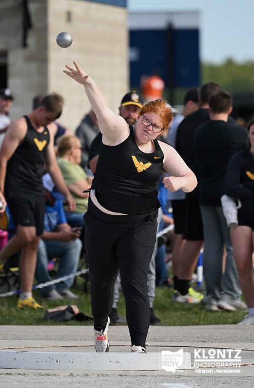 Female shot-put athlete mid-throw on the throwing circle, shot in the air, surrounded by spectators in the background.