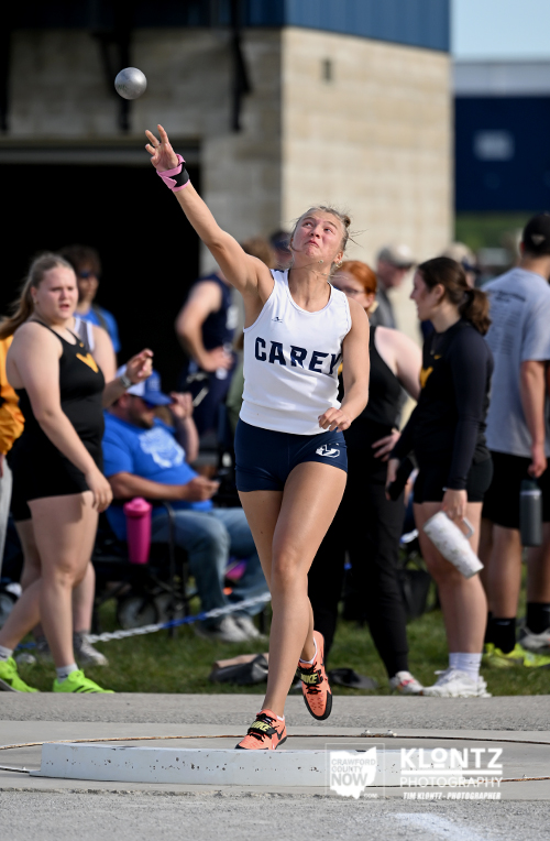 Female athlete in a CARE jersey releases a shot put during a track meet, with spectators in the background.