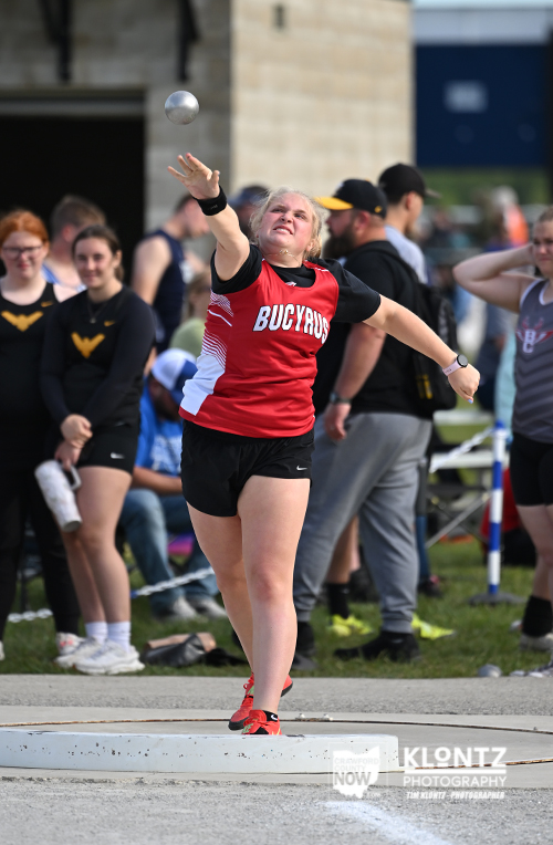 Female athlete in a red Bucyrus jersey is throwing a hammer during a track and field event, with spectators watching in the background.