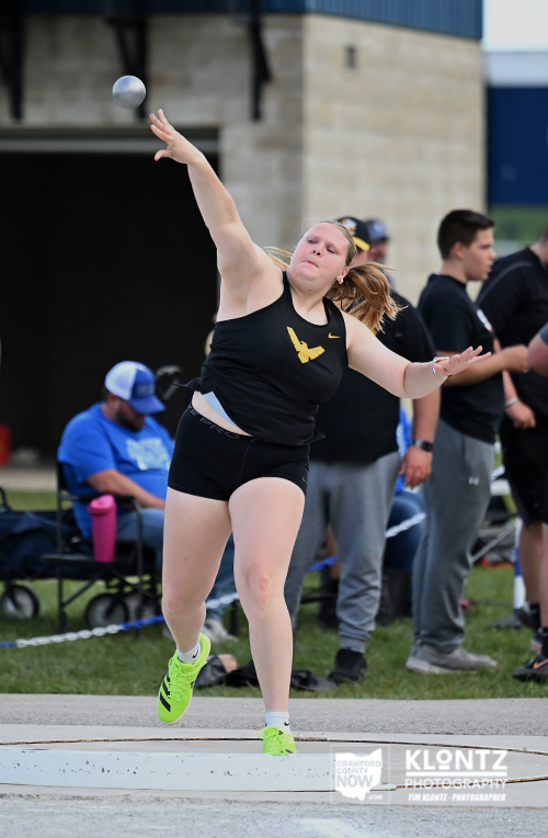 Female shot-put athlete in black uniform releasing a shot put on the throwing circle, with spectators in the background.