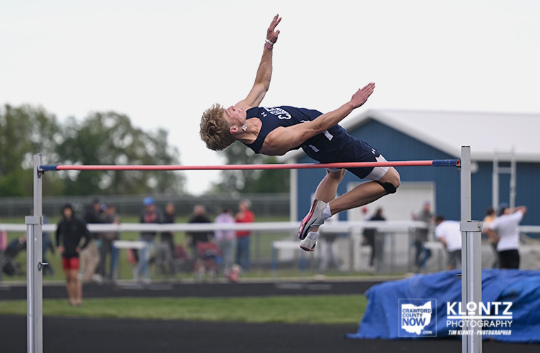 Male athlete in a navy uniform clears a high jump bar mid-air on an outdoor track field, with spectators in the background and a blue building nearby.
