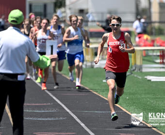 Male sprinter in a red jersey leads a track race as runners follow on the lane, with a race official in a green cap signaling from the left.
