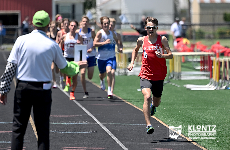 Male sprinter in a red jersey leads a track race as runners follow on the lane, with a race official in a green cap signaling from the left.