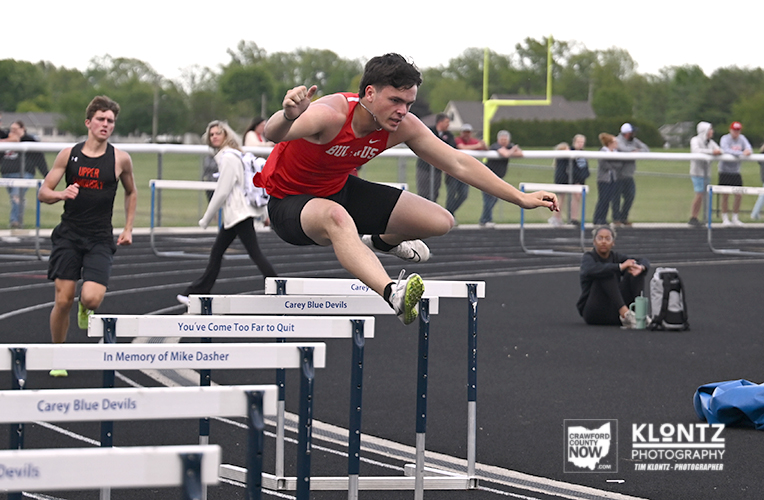 Athlete in red jersey clears a hurdle during a track race on an outdoor field, mid-air with a focused expression as teammates and spectators watch nearby.
