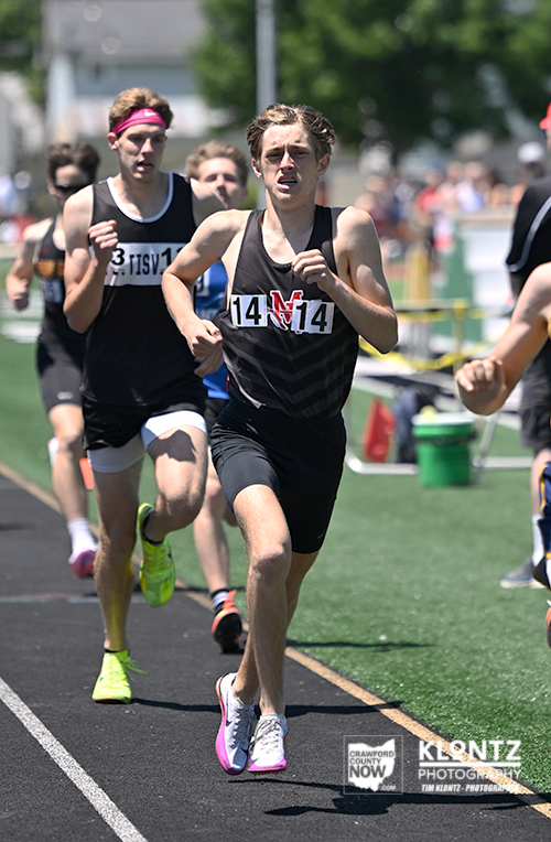 Young male sprinter in a black racing singlet with bib 14 leads a outdoor track race, other runners behind him on the track.