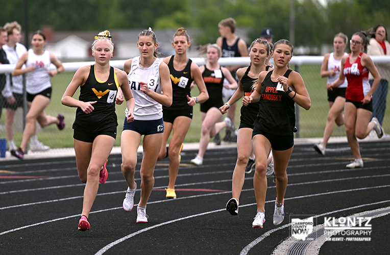 Female high school runners sprint on an outdoor track during a race; front runners wear black and white uniforms.