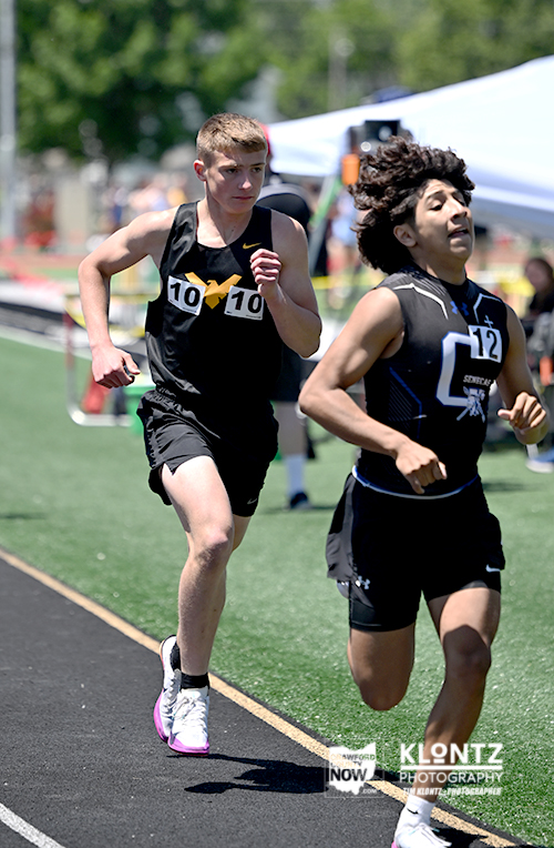 Two male sprinters race side by side on a track, wearing bib numbers 10 and 12, concentrating as they push toward the finish line.