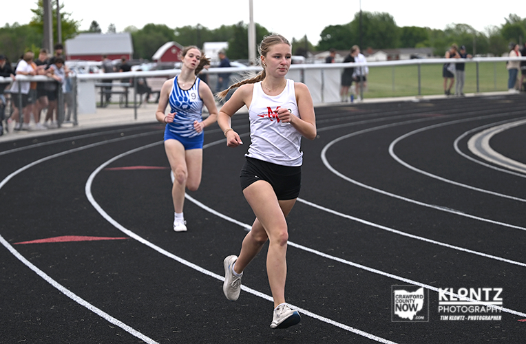 Two female sprinters race on a curved track; the lead runner wears a white tank top and black shorts, while a blue-uniformed runner follows behind as spectators watch from the sidelines.