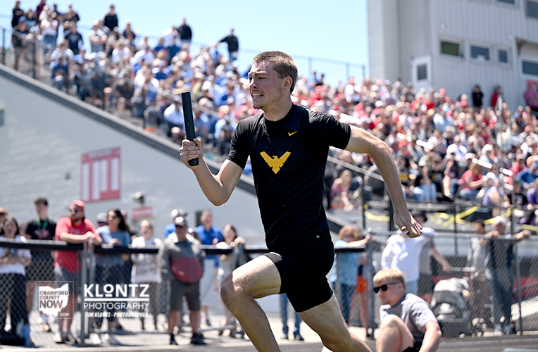 Male sprinter holding a baton while running on a track in a stadium, with a crowded stands audience in the background for a relay race.