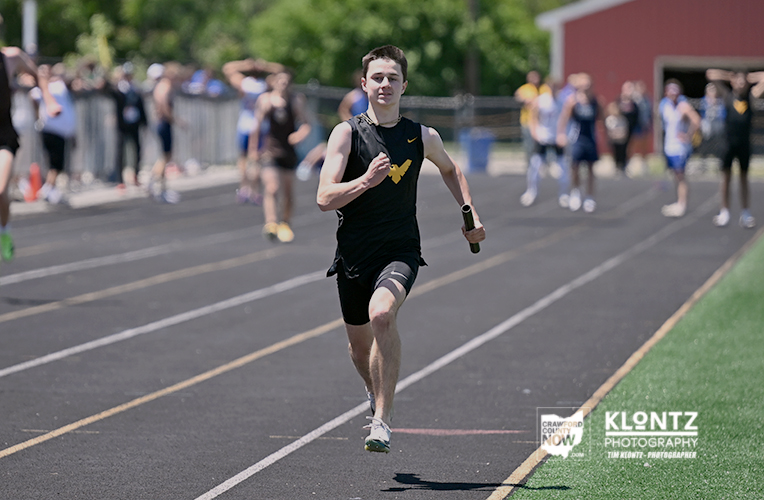 Male sprinter in a black uniform runs toward the camera on an outdoor track, holding a baton with spectators in the background.