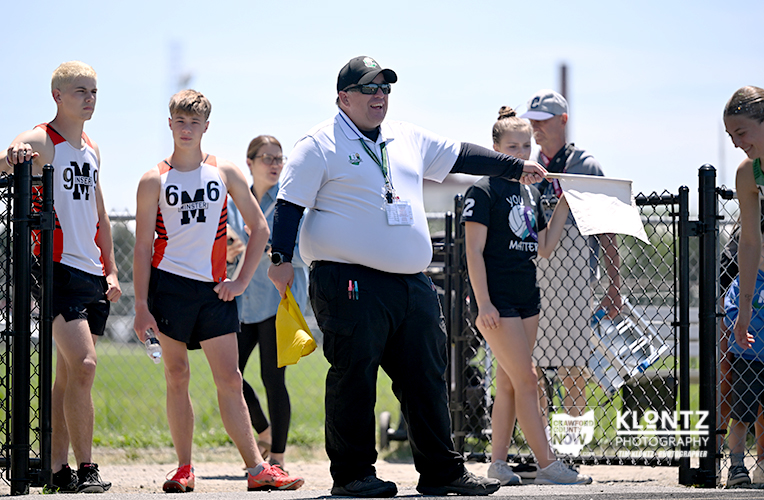 Coach in a white shirt and cap speaks to young track athletes beside a fence on a sunny day, before a race.