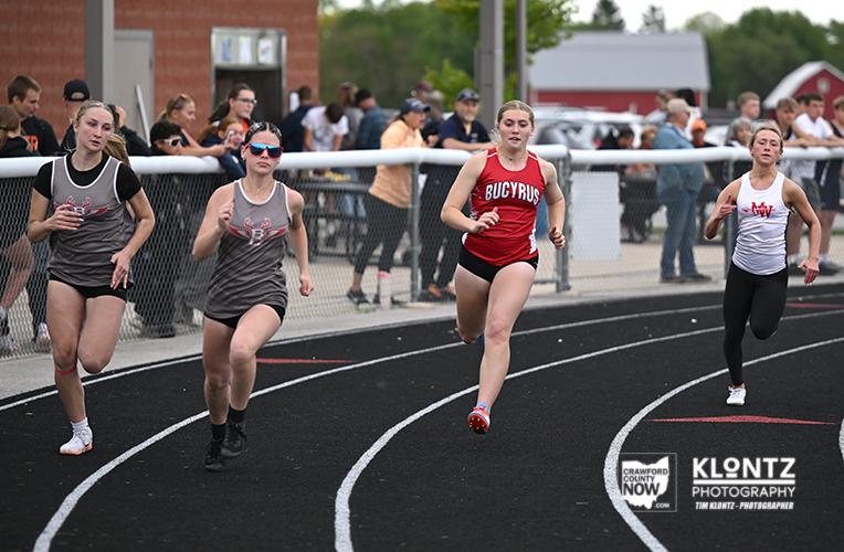 Four female sprinters race on a curved outdoor track as spectators watch from behind a fence; the lead runner wears a red Bucyrus uniform.