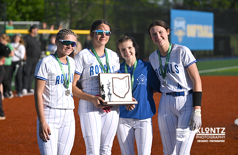 Four Royals softball players in white pinstripe uniforms pose with medals and a trophy on a softball field.