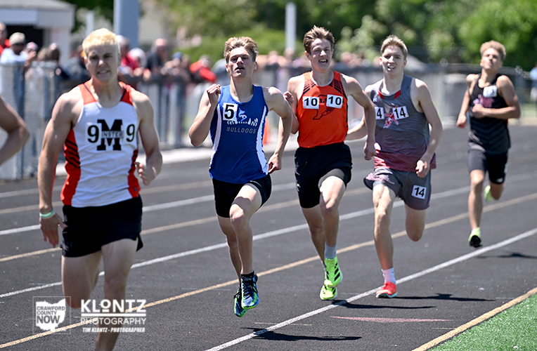 Four male sprinters race on an outdoor track, wearing numbered bibs (9, 15, 10, 14) and colorful uniforms, while spectators line the stands nearby.
