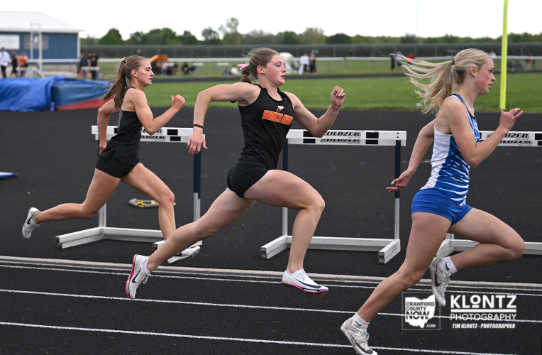 Three female sprinters race on a track with hurdles in the background during a competition at an outdoor track.