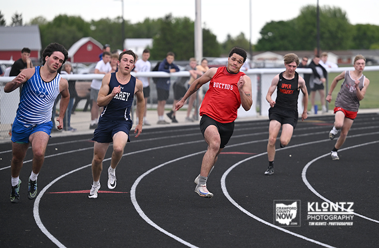 Five male sprinters race on a track, leaning into the bend as they push toward the finish while spectators watch in the background.