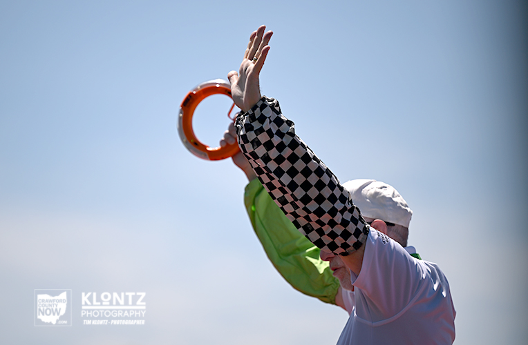 Person in a white shirt and checkered sleeve raises an orange ring against a clear blue sky, participating in a ring toss game outdoors.