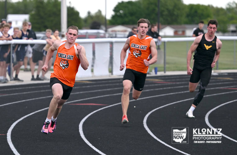 Three male sprinters racing on a track; two wear orange 'Seneca East' uniforms while a black-clad runner trails behind, with a crowd in the background.