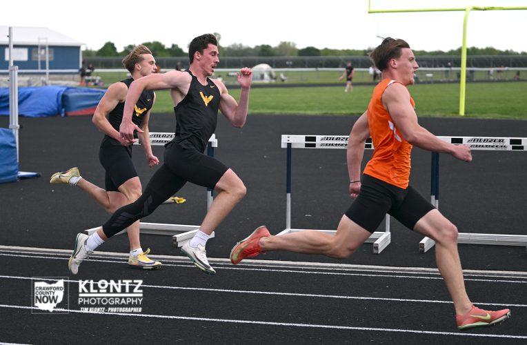 Three male sprinters in a track race; lead runner in an orange singlet ahead of two rivals in black, with hurdles visible on the track.